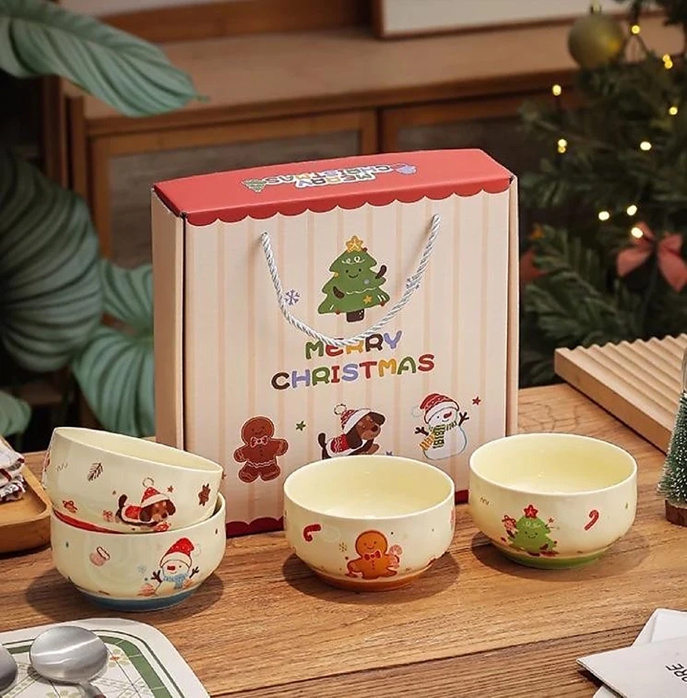 Set bốn bát sứ Giáng sinh họa tiết người tuyết và bánh quy gừng đặt cạnh hộp quà Merry Christmas có quai xách