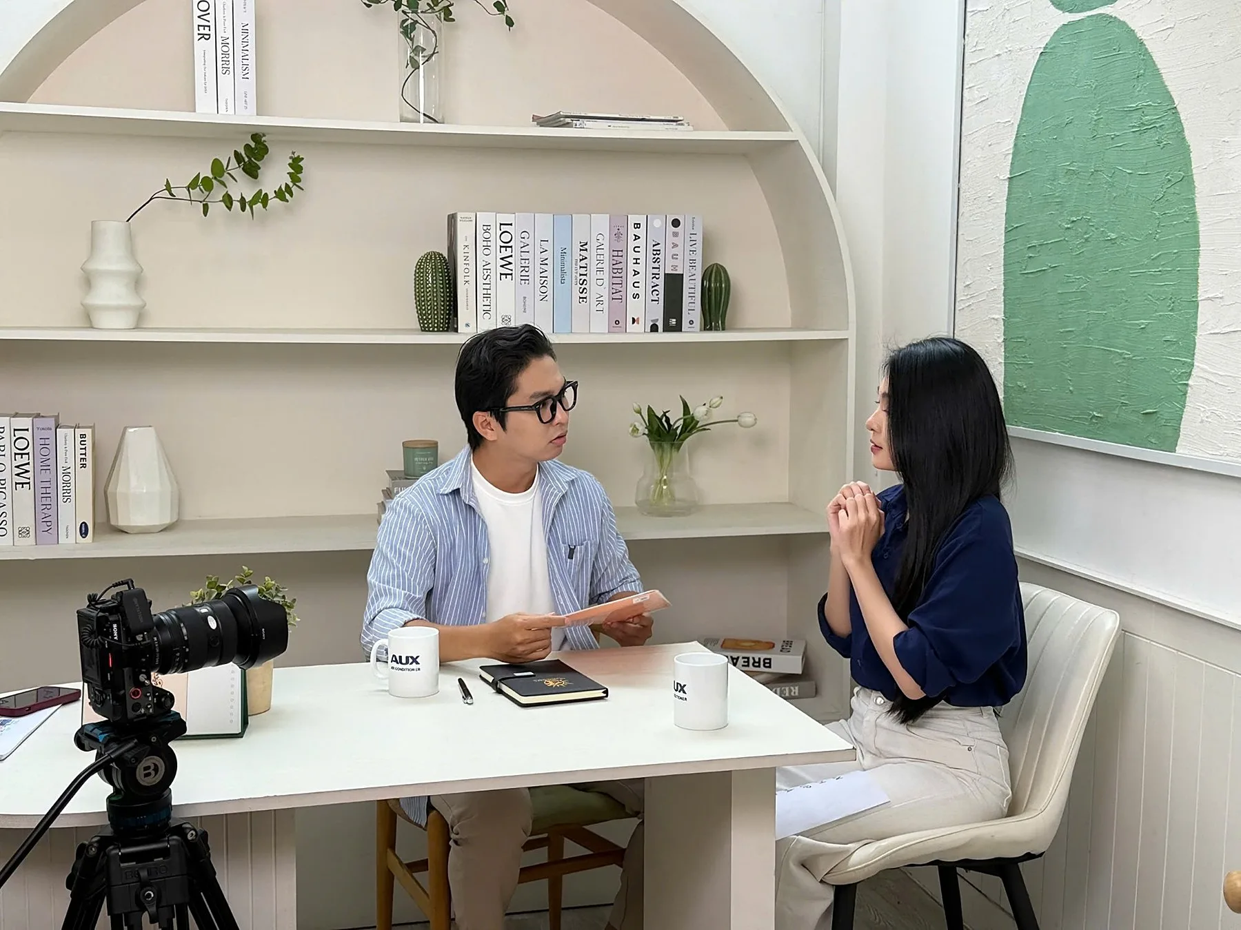 Two actors in conversation in a living room set with AUX mugs on the table and a Sony camera filming